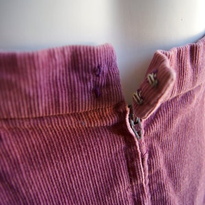 May include: Close-up of a pink corduroy skirt with a zipper and a button closure. The fabric has a subtle texture and a slightly faded appearance.