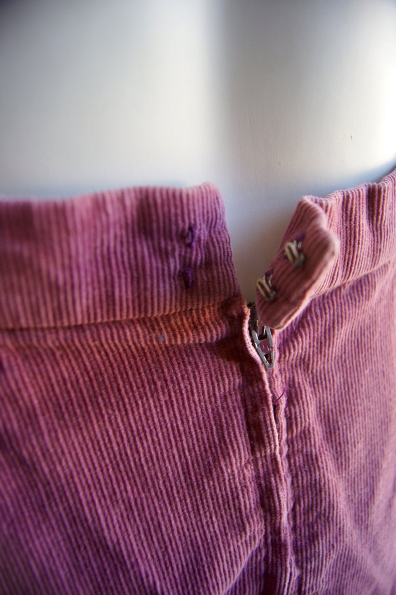 May include: Close-up of a pink corduroy skirt with a zipper and a button closure. The fabric has a subtle texture and a slightly faded appearance.
