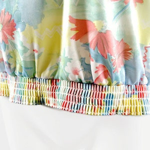 May include: A floral print top with a gathered elastic waistband. The fabric features a pattern of pink, red, and white flowers on a light blue and yellow background. The elastic band is a rainbow of colors.