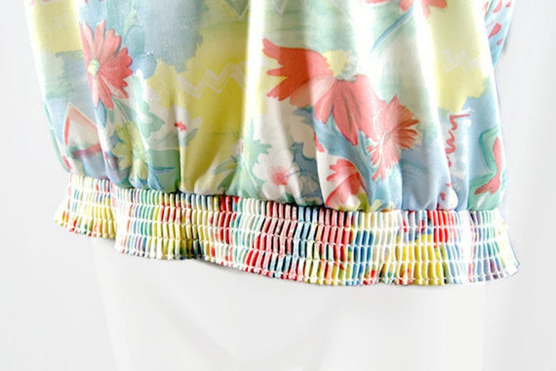 May include: A floral print top with a gathered elastic waistband. The fabric features a pattern of pink, red, and white flowers on a light blue and yellow background. The elastic band is a rainbow of colors.