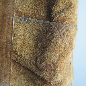 May include: Close-up of a brown suede jacket with a stitched pocket on the front. The pocket has a decorative design.