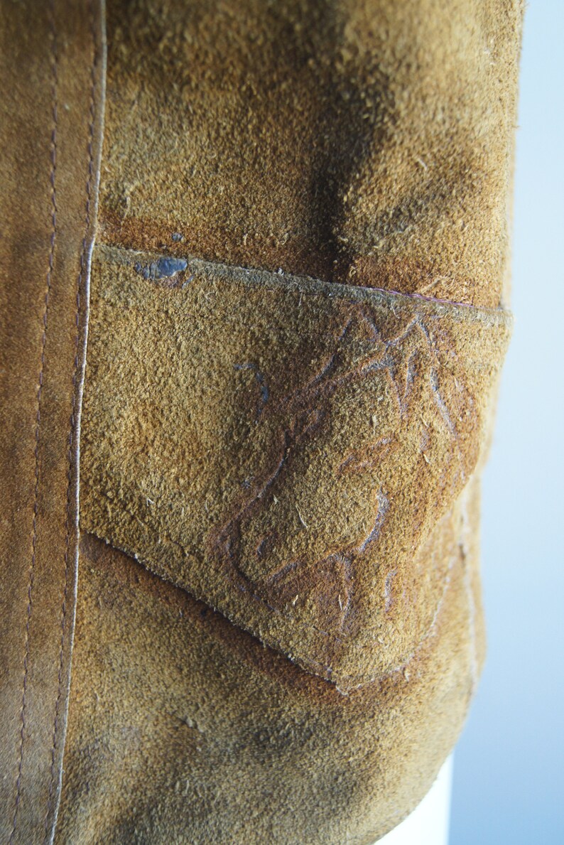 May include: Close-up of a brown suede jacket with a stitched pocket on the front. The pocket has a decorative design.