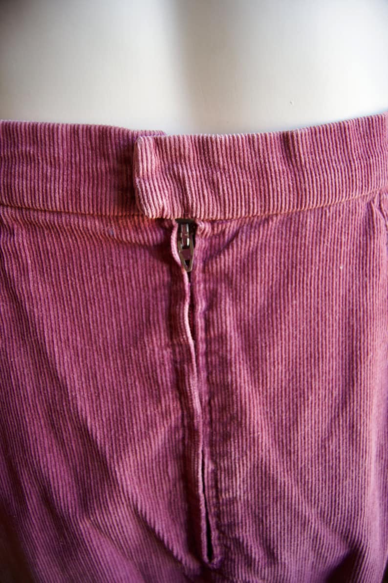 May include: A close-up of the back of a pink corduroy skirt with a zipper closure.