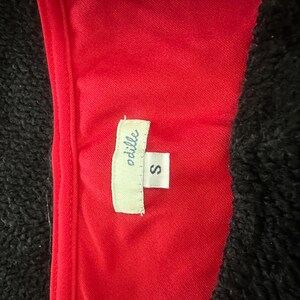 May include: Close-up of a red garment with a tag that reads "odille" and the size "S". The garment is set against a black, textured background. The tag is white with black text.