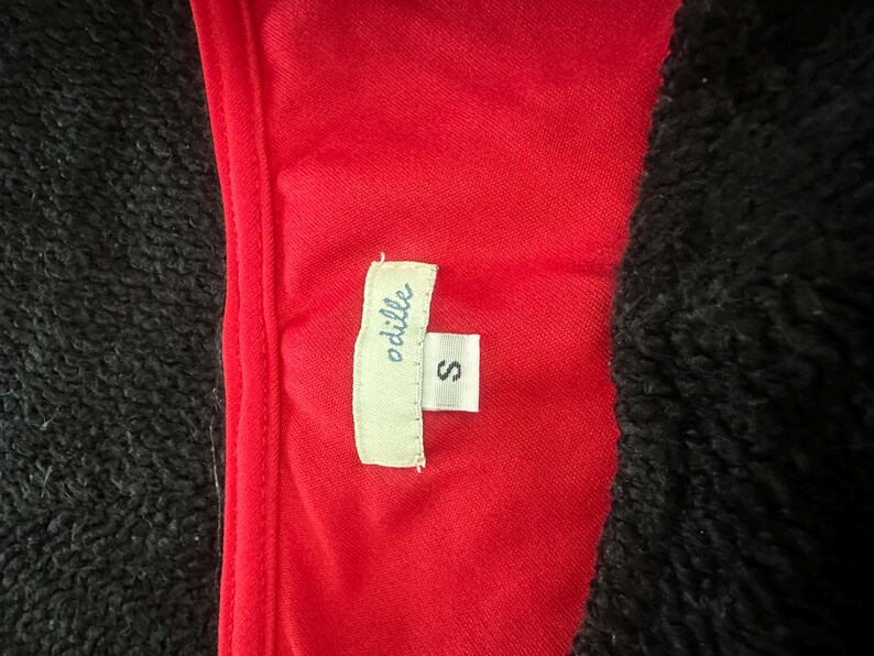 May include: Close-up of a red garment with a tag that reads "odille" and the size "S". The garment is set against a black, textured background. The tag is white with black text.