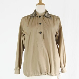 May include: A tan long-sleeved shirt with a plaid collar and black buttons. The shirt has a drawstring at the bottom.