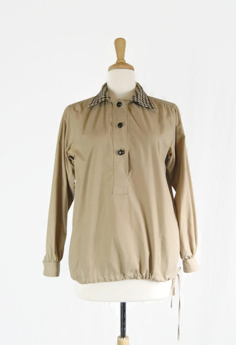 May include: A tan long-sleeved shirt with a plaid collar and black buttons. The shirt has a drawstring at the bottom.