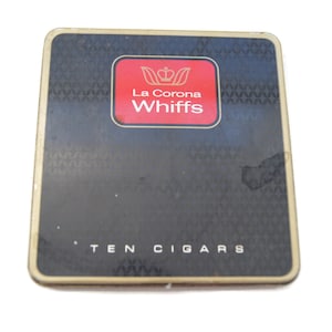 Puede incluir: Un paquete de cigarrillos negro y dorado con un cuadrado rojo que dice "La Corona Whiffs" y una corona dorada. El texto "TEN CIGARS" está impreso en la parte inferior del paquete.