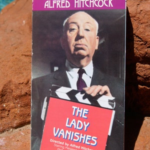 May include: A VHS tape cover featuring Alfred Hitchcock holding a clapperboard. The cover is purple and white with the title "The Lady Vanishes" in blue and white text. The text "Directed by Alfred Hitchcock" is also included.