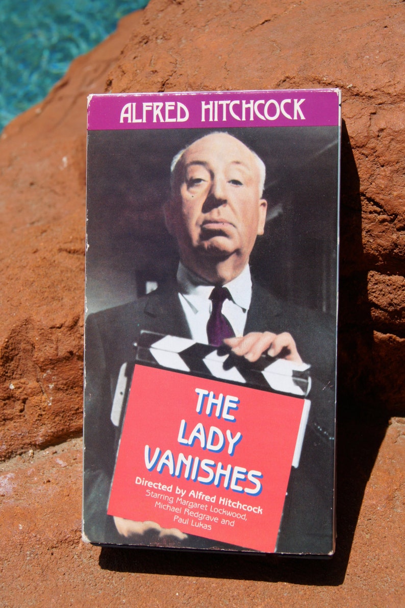 May include: A VHS tape cover featuring Alfred Hitchcock holding a clapperboard. The cover is purple and white with the title "The Lady Vanishes" in blue and white text. The text "Directed by Alfred Hitchcock" is also included.