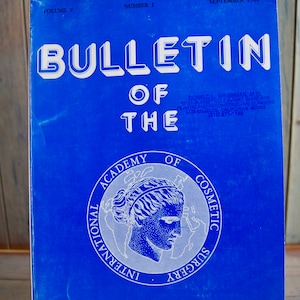 May include: A blue cover of the Bulletin of the International Academy of Cosmetic Surgery, Volume V, Number 1, September 1980. The cover features a white logo with a woman's head and the words "International Academy of Cosmetic Surgery".