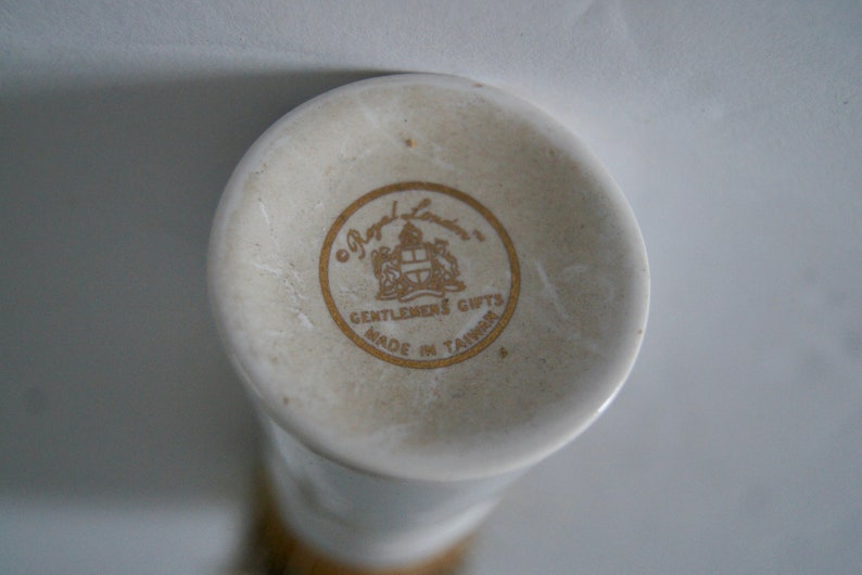 May include: Close-up of a white ceramic bottom of a vase with a gold logo that reads "Royal London, Gentlemen's Gifts, Made in Taiwan."