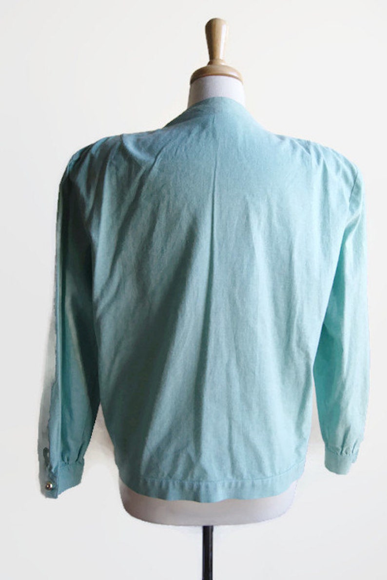 Pode incluir: Uma blusa azul claro de manga comprida com fecho de bot&otilde;es na frente e manga ligeiramente bufante.