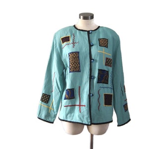 Puede incluir: Una chaqueta azul claro con ribete negro y bordado geométrico de colores. La chaqueta tiene cierre de botones y mangas largas.