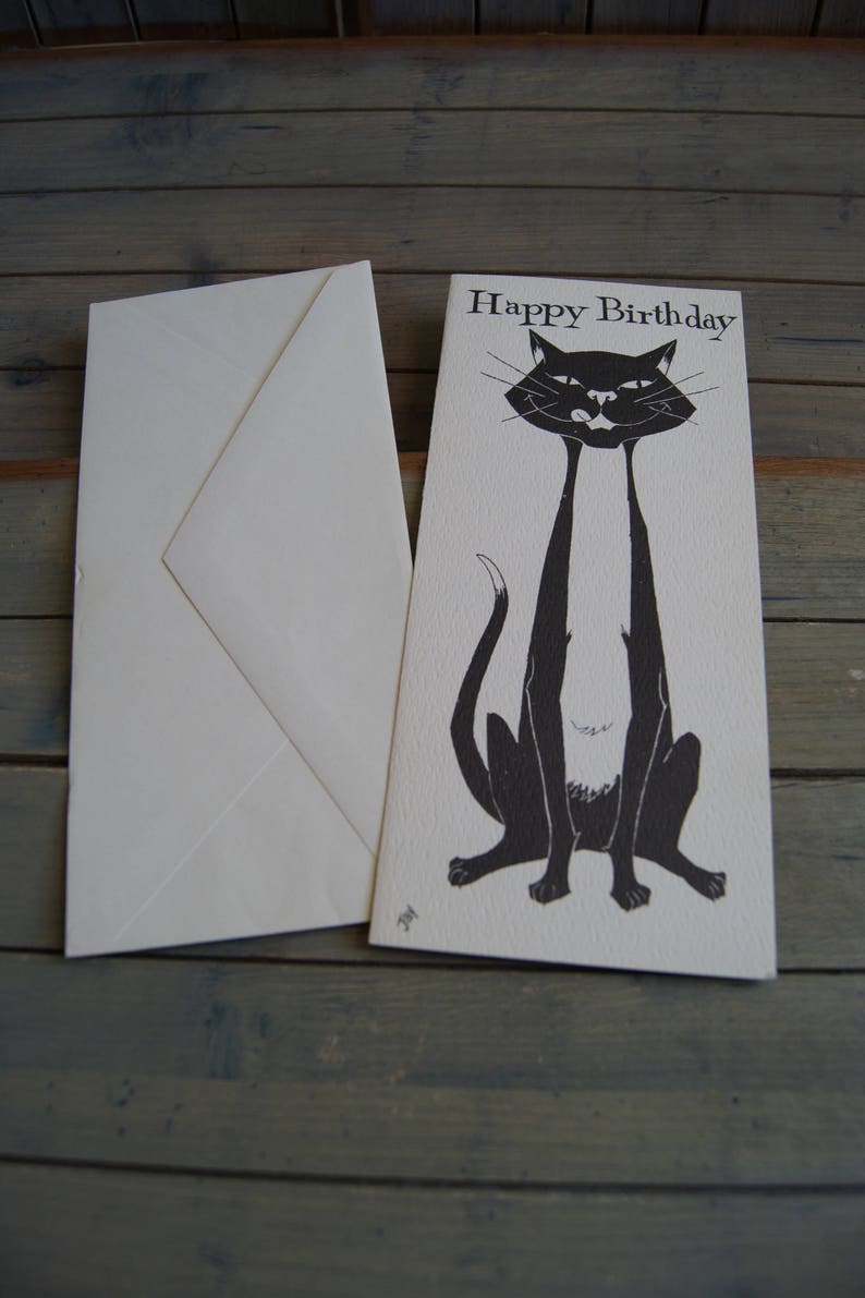 Op de afbeelding: Een witte kaart met zwarte tekst die "Happy Birthday" zegt. Een zwart-witte illustratie van een kat met lange benen staat op de kaart.