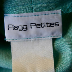 Pode incluir: Etiqueta branca costurada em uma pe&ccedil;a de roupa de tecido turquesa com o texto "Flagg Petites" em preto.