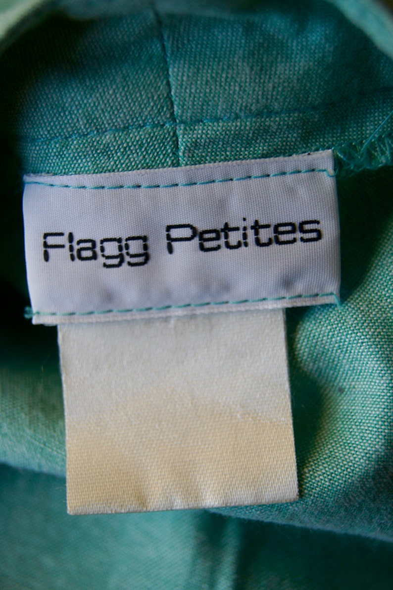 Pode incluir: Etiqueta branca costurada em uma pe&ccedil;a de roupa de tecido turquesa com o texto "Flagg Petites" em preto.