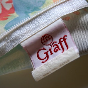 May include: Close-up of a fabric with a white zipper and a tag. The tag has the word "Graff" in red script and a globe logo. The fabric has a floral pattern with colors like pink, blue, and yellow.