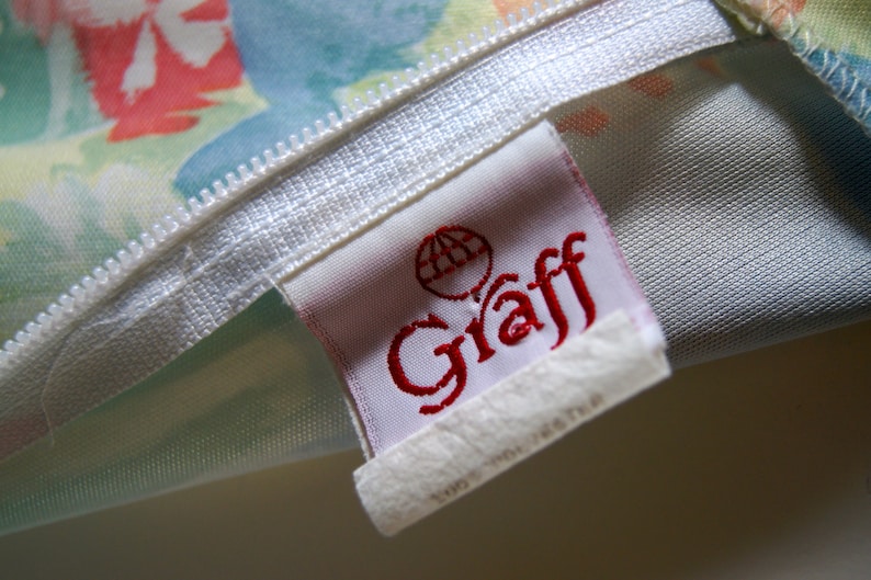 May include: Close-up of a fabric with a white zipper and a tag. The tag has the word "Graff" in red script and a globe logo. The fabric has a floral pattern with colors like pink, blue, and yellow.