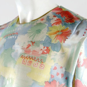 May include: A vintage floral print blouse featuring a watercolor-style design. The blouse showcases a pastel color palette with light blue, yellow, and green hues, accented by red and pink floral patterns. The fabric appears soft and lightweight.