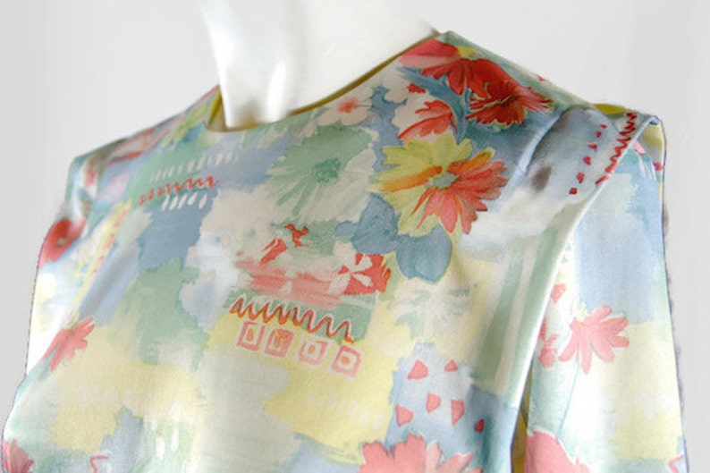 May include: A vintage floral print blouse featuring a watercolor-style design. The blouse showcases a pastel color palette with light blue, yellow, and green hues, accented by red and pink floral patterns. The fabric appears soft and lightweight.