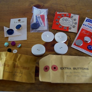 Op de afbeelding: Een assortiment knopen in verschillende kleuren en maten, waaronder blauw, turquoise, paars en wit. Sommige knopen zijn verpakt in plastic zakjes, terwijl andere los liggen. De knopen zijn gelabeld met "Extra Buttons" en "La Mode".