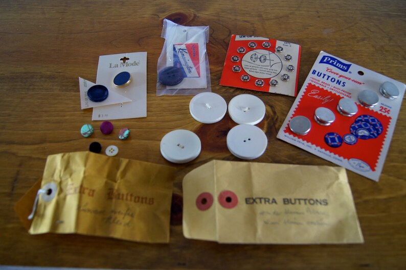 Op de afbeelding: Een assortiment knopen in verschillende kleuren en maten, waaronder blauw, turquoise, paars en wit. Sommige knopen zijn verpakt in plastic zakjes, terwijl andere los liggen. De knopen zijn gelabeld met "Extra Buttons" en "La Mode".