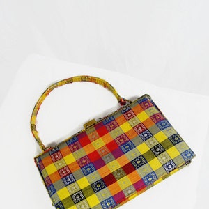 Puede incluir: Un bolso vintage con un colorido estampado a cuadros en amarillo, naranja, rojo, morado y azul. El bolso tiene un cierre dorado y un asa estampada a juego. El bolso es rectangular y parece estar hecho de tela.