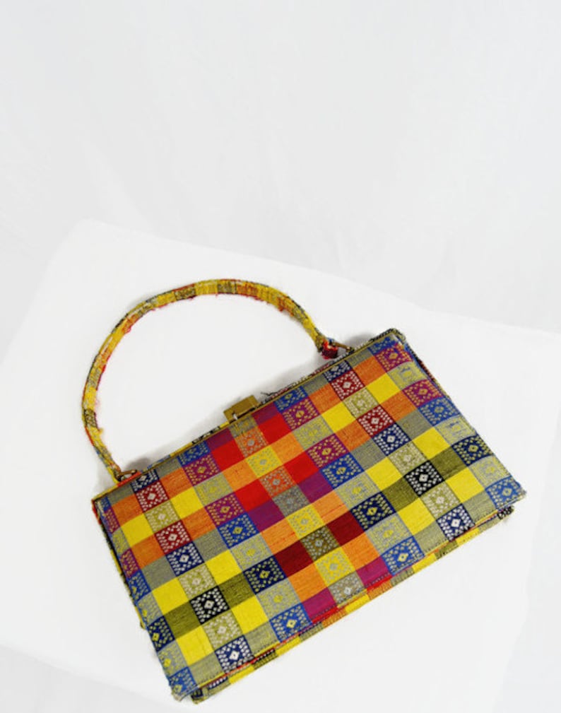 Puede incluir: Un bolso vintage con un colorido estampado a cuadros en amarillo, naranja, rojo, morado y azul. El bolso tiene un cierre dorado y un asa estampada a juego. El bolso es rectangular y parece estar hecho de tela.