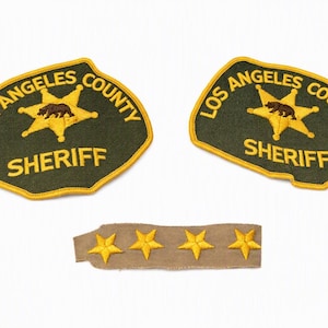 May include: Two dark green Los Angeles County Sheriff patches with yellow embroidered borders, text, and a central star featuring a brown bear. Below them is a tan rectangular fabric strip with four embroidered yellow stars in a row.