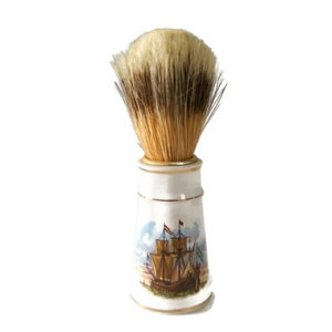 May include: A white ceramic shaving brush with a gold trim and a painted image of a ship. The brush has a brown and white bristle head.