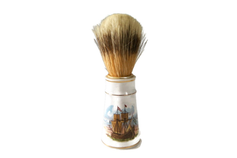 May include: A white ceramic shaving brush with a gold trim and a painted image of a ship. The brush has a brown and white bristle head.