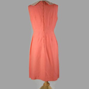 May include: A sleeveless coral dress with a white lace collar. The dress is on a wooden mannequin and has a back zipper.
