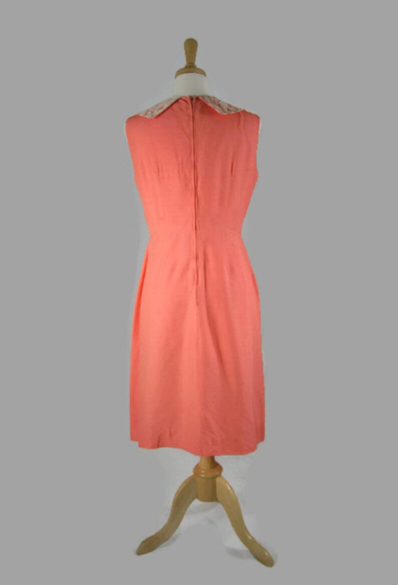 May include: A sleeveless coral dress with a white lace collar. The dress is on a wooden mannequin and has a back zipper.