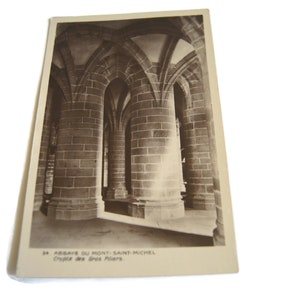 May include: Black and white photograph postcard of the interior of the Abbey of Mont-Saint-Michel. The photo shows the stone columns and arches of the crypt of the Gros Piliers.