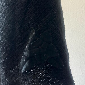 May include: Close-up of a black fabric with a textured surface. A triangular, quilted patch is sewn onto the fabric. The patch has a geometric design and is also black.
