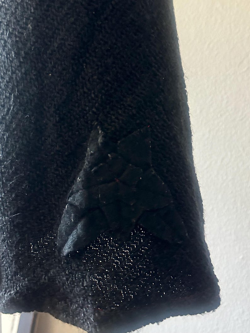 May include: Close-up of a black fabric with a textured surface. A triangular, quilted patch is sewn onto the fabric. The patch has a geometric design and is also black.