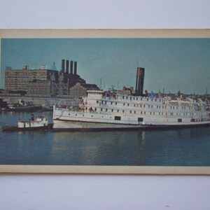 Könnte beinhalten: Eine Vintage-Postkarte mit einem weißen Dampfschiff mit einem roten Schornstein, das an einem Pier angelegt ist. Das Schiff ist von Wasser umgeben und im Hintergrund ist eine Stadt zu sehen.