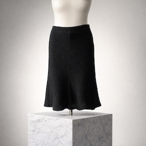 May include: A black, textured skirt displayed on a mannequin. The skirt has a flared hem and a fitted waist. The fabric appears to be a woven material. The skirt is displayed on a white mannequin torso and a marble block.
