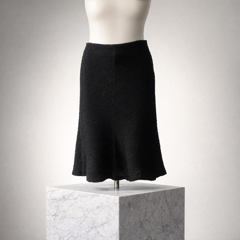May include: A black, textured skirt displayed on a mannequin. The skirt has a flared hem and a fitted waist. The fabric appears to be a woven material. The skirt is displayed on a white mannequin torso and a marble block.