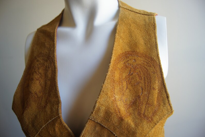 May include: A brown suede vest with a deep V-neck and decorative stitching. The vest has a unique design with two circular patterns on the front.