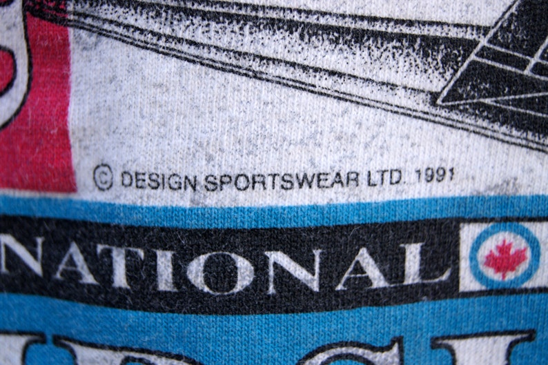 May include: A white t-shirt with a blue and black graphic of a hockey player and the text "National Hockey League". The shirt also has the text "&copy; DESIGN SPORTSWEAR LTD. 1991" printed on it.