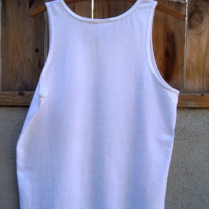 May include: A white sleeveless tank top hanging on a wooden hanger. The tank top is plain and has no visible graphics or designs.