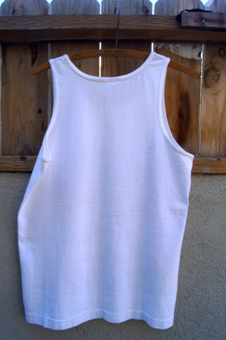 May include: A white sleeveless tank top hanging on a wooden hanger. The tank top is plain and has no visible graphics or designs.