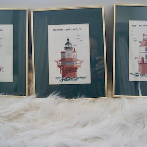 May include: Three framed cross-stitch artworks featuring lighthouse illustrations. Each piece has a dark green mat and a thin gold frame. The left depicts Thimble Shoal Light, VA; the center, Solomons Lump Light, MD; and the right, Point No Point Light, MD.