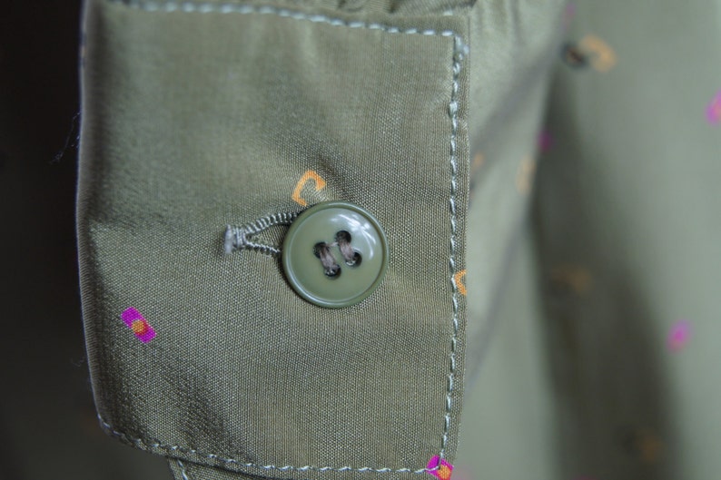 May include: A close-up of a green button with four holes on a green fabric with a white stitch around the edge.