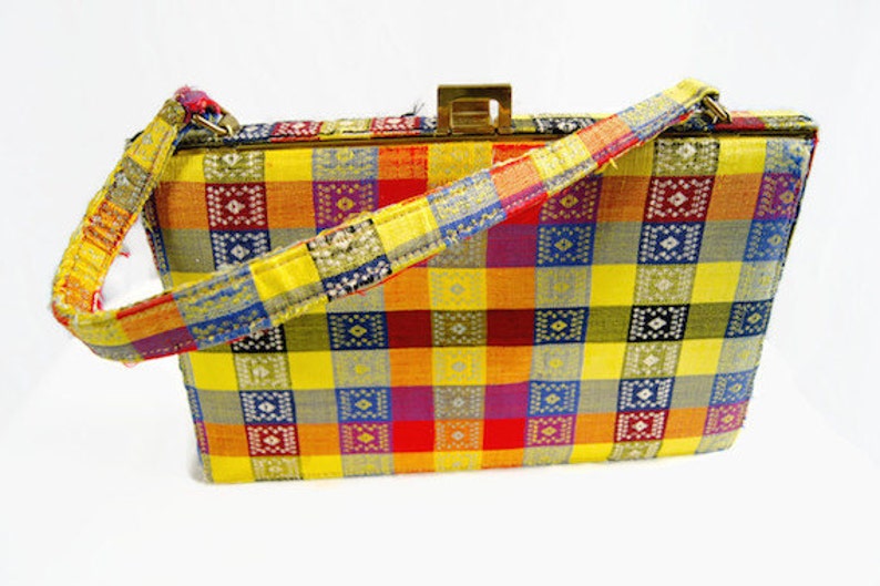 Puede incluir: Un bolso vintage con un colorido estampado a cuadros en amarillo, naranja, rojo, morado, azul y verde. El bolso tiene forma rectangular, un cierre dorado y un asa con estampado a juego.