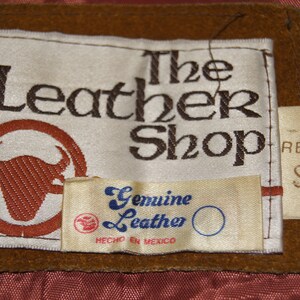 May include: A brown leather label with white text that reads "The Leather Shop" with a brown bull's head logo. A smaller white label below reads "Genuine Leather" with blue text and "Hecho en Mexico". A smaller white label on the right side reads "Reg S".