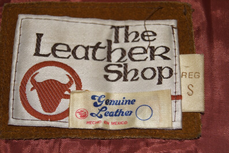 May include: A brown leather label with white text that reads "The Leather Shop" with a brown bull's head logo. A smaller white label below reads "Genuine Leather" with blue text and "Hecho en Mexico". A smaller white label on the right side reads "Reg S".