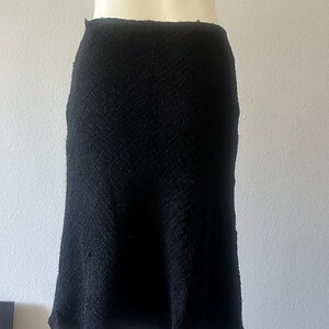 May include: A black, textured skirt with a slight A-line shape. The skirt appears to be made of a woven fabric, and it is displayed on a mannequin. The skirt's hemline falls below the knees.